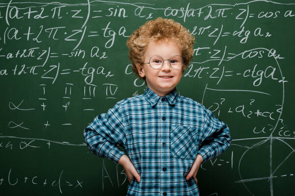 happy kid in glasses standing with hands on hips near chalkboard 