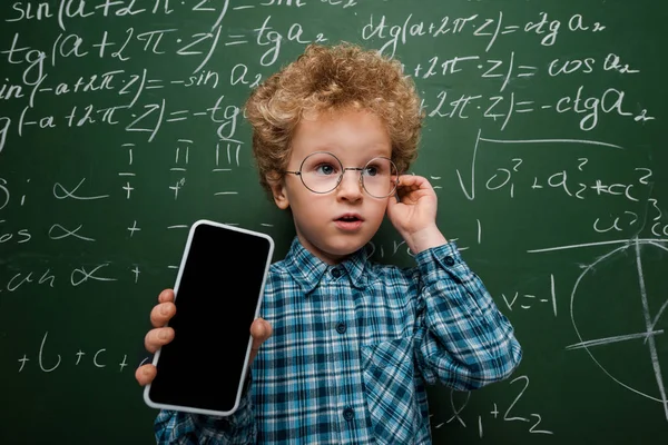 戴眼镜的聪明孩子拿着装有空白屏幕的智能手机 靠近黑板 附有数学公式 — 图库照片