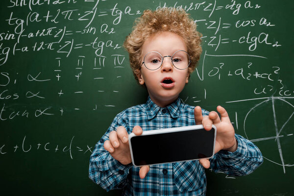 smart kid in glasses holding smartphone with blank screen near chalkboard with mathematical formulas 