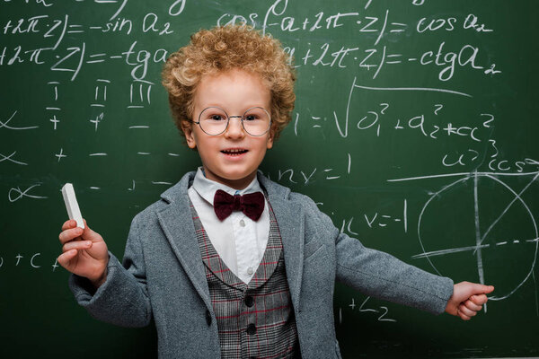 happy kid in suit with bow tie holding chalk near chalkboard with mathematical formulas 