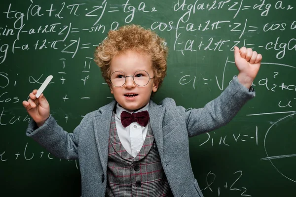 聪明的孩子穿着西装 带着弓形领带 在黑板附近拿着粉笔 附有数学公式 — 图库照片