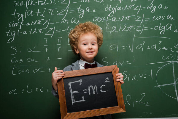 happy kid in suit and bow tie holding small blackboard with formula near chalkboard 