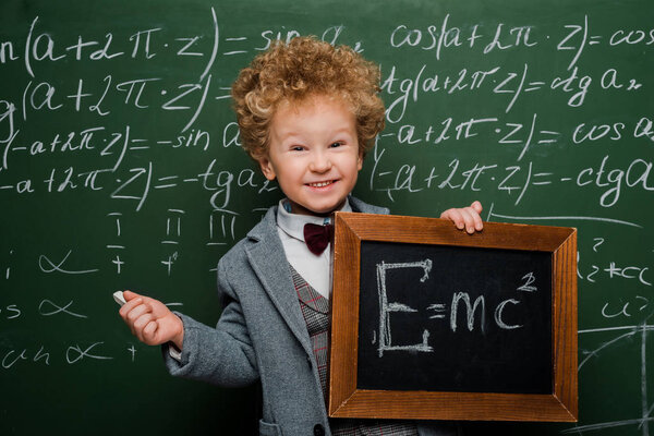 smiling and smart kid in suit and bow tie holding small blackboard with formula near chalkboard 