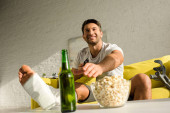 Szelektív fókusz mosolygós férfi törött lábbal tv-nézés közel sörösüveg és popcorn a nappaliban