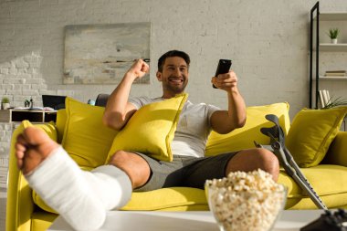 Selective focus of cheerful man with broken leg watching tv near popcorn on table in living room