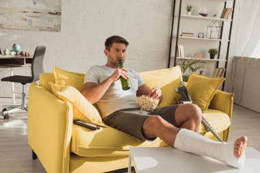 Handsome man with broken leg holding popcorn and beer while watching tv in living room