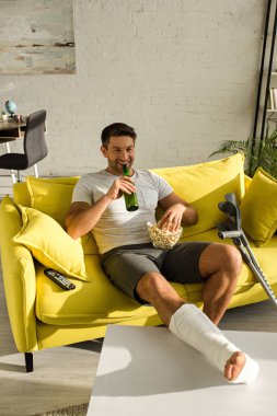 Smiling man with broken leg drinking beer and holding popcorn while watching movie on couch