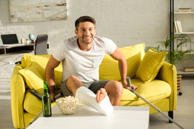 Smiling man with broken leg looking at camera near beer bottle and popcorn on coffee table in living room