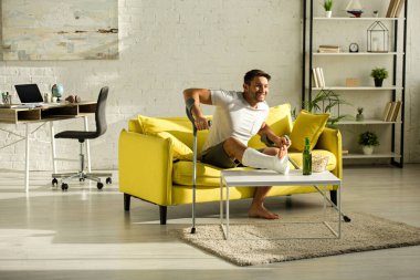 Smiling man with broken leg and crutches and watching tv near beer and popcorn in living room