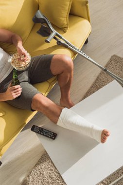 Top view of man with broken leg on coffee table drinking beer and eating popcorn on sofa at home