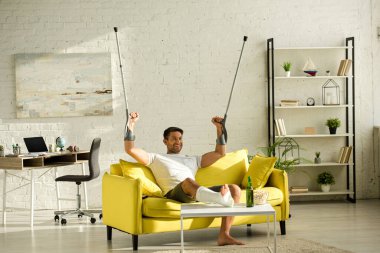 Cheerful man with broken leg and crutches watching tv near popcorn and beer in living room