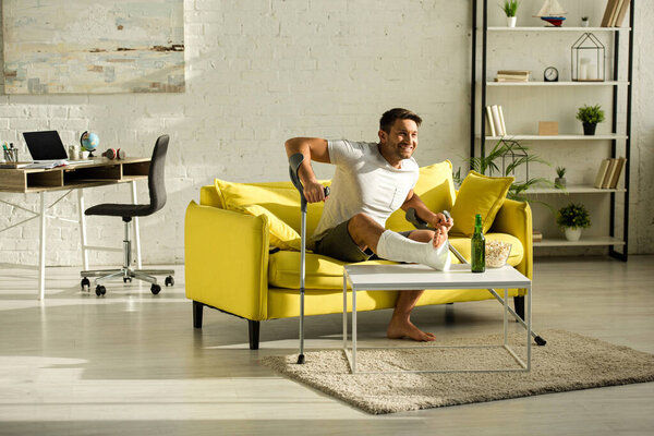 Улыбающийся мужчина со сломанной ногой и костылями и смотрящий телевизор возле пива и попкорна в гостиной
