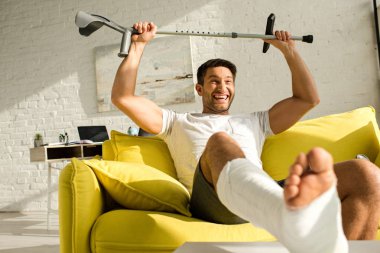 Selective focus of positive man with broken leg raising crutch while sitting on couch at home