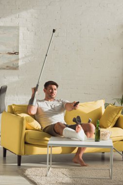 Angry man with broken leg watching tv near beer bottle and popcorn on coffee table at home