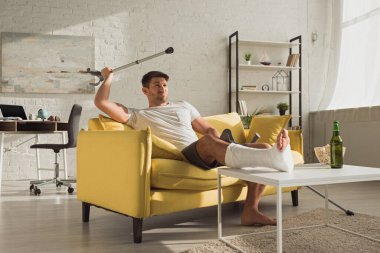 Handsome man with broken leg raising crutch near beer and popcorn on coffee table in living room