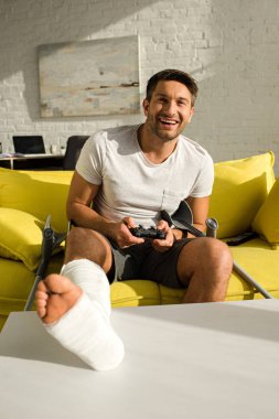 KYIV, UKRAINE - JANUARY 21, 2020: Selective focus of smiling man with broken leg holding joystick and looking at camera on couch