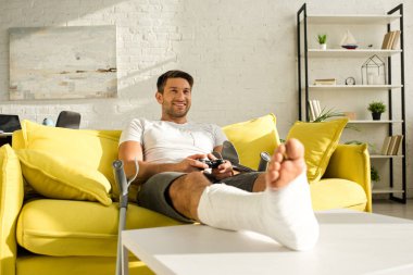 KYIV, UKRAINE - JANUARY 21, 2020: Handsome smiling man with broken leg on coffee table playing video game in living room