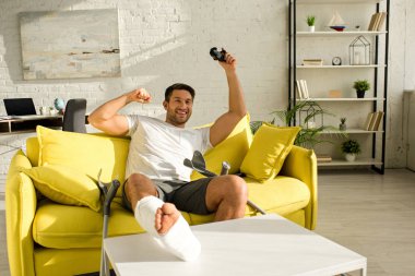 KYIV, UKRAINE - JANUARY 21, 2020: Cheerful man with broken leg smiling while holding joystick on couch at home