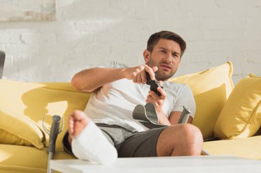 KYIV, UKRAINE - JANUARY 21, 2020: Selective focus of handsome man with broken leg playing video game near crutches on sofa 