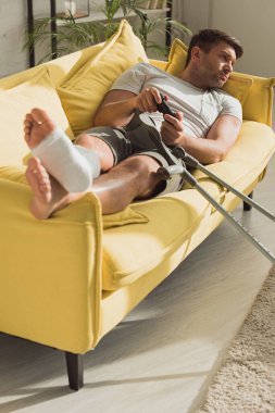KYIV, UKRAINE - JANUARY 21, 2020: Selective focus of man with broken leg playing video game on sofa at home