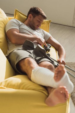 KYIV, UKRAINE - JANUARY 21, 2020: Handsome man with broken leg sleeping near joystick and crutches on couch  