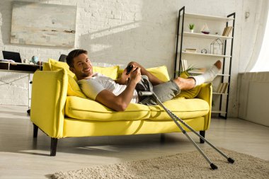 KYIV, UKRAINE - JANUARY 21, 2020: Handsome man with broken leg smiling at camera and holding joystick on sofa at home