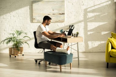 Side view of freelancer with broken leg working at laptop in living room