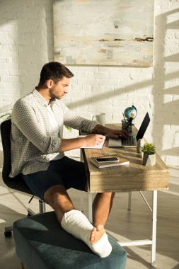 Side view of freelancer with broken leg using laptop near notebooks and smartphone on table