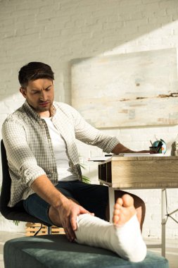Selective focus of handsome man touching broken leg while sitting at table in living room