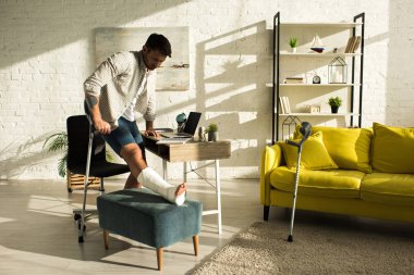 Handsome man with broken leg on ottoman holding crutch in living room
