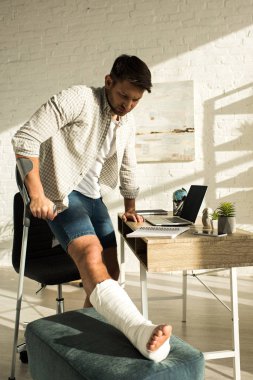 Man with broken leg standing near table with laptop and notebooks