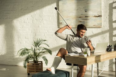 Smiling man with broken leg holding crutch and smartphone white sitting at table with laptop