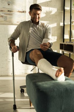 Selective focus of smiling man with broken leg using smartphone in living room