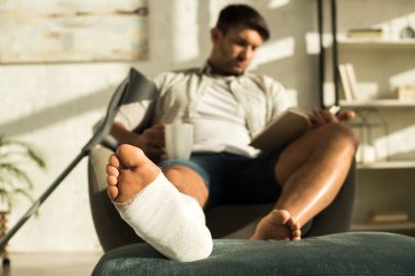 Selective focus of man with leg in plaster bandage holding cup and reading book in armchair 