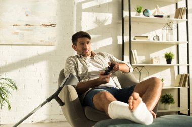 KYIV, UKRAINE - JANUARY 21, 2020: Handsome man with broken leg on ottoman playing video game at home
