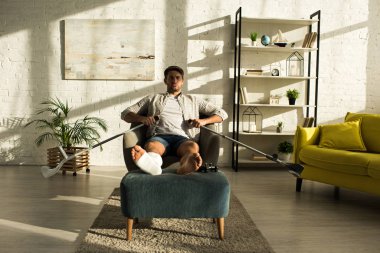 KYIV, UKRAINE - JANUARY 21, 2020: Handsome man with broken leg on ottoman holding crutches while sitting in armchair in living room