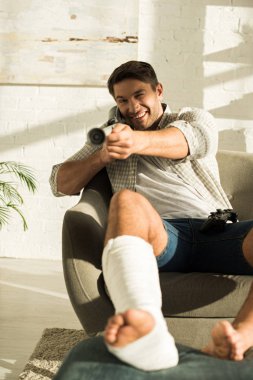 KYIV, UKRAINE - JANUARY 21, 2020: Selective focus of smiling man with broken leg holding crutch and looking at camera at home