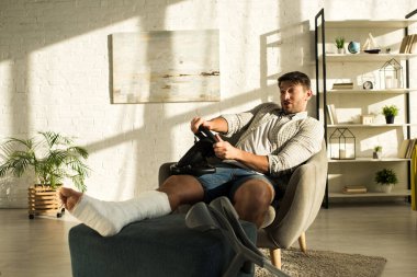 Handsome man with broken leg playing with steering wheel in video game at home