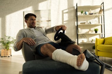 Handsome man with broken leg playing video game with steering wheel in living room
