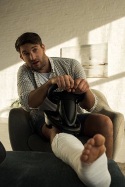 Selective focus of man with leg in plaster bandage holding playing steering wheel in living room