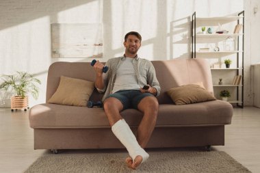 Smiling man with broken leg watching tv and holding dumbbell on sofa at home
