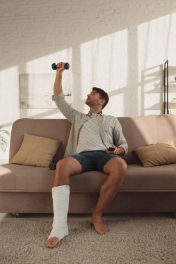 Handsome man with leg in plaster bandage training with dumbbell and holding remote controller on couch