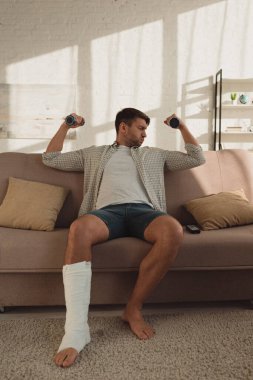 Handsome man with broken leg training with dumbbells on couch in living room