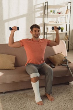 Smiling man with broken leg exercising with dumbbells near crutches on couch