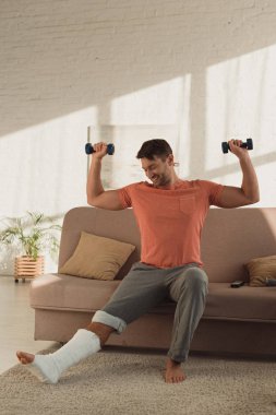 Smiling man looking at broken leg while training with dumbbells on sofa