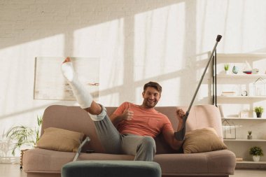 Handsome man with raised broken leg and crutch showing approval sign at camera on couch
