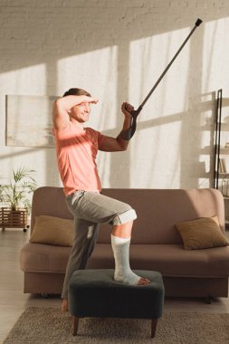 Man with broken leg on ottoman smiling away and holding crutch in sunlit living room 
