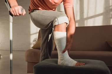 Cropped view of man touching leg in plaster bandage on ottoman and holding crutch in living room