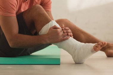 Cropped view of man holding broken leg and sitting on fitness mat on floor clipart