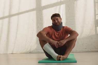 Man with neck brace and broken leg smiling at camera on fitness mat on floor